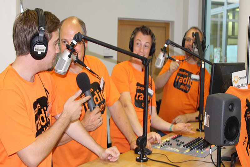 Vier Männer in orangenen Shirts sprechen angeregt vor zwei Mikrofonen.