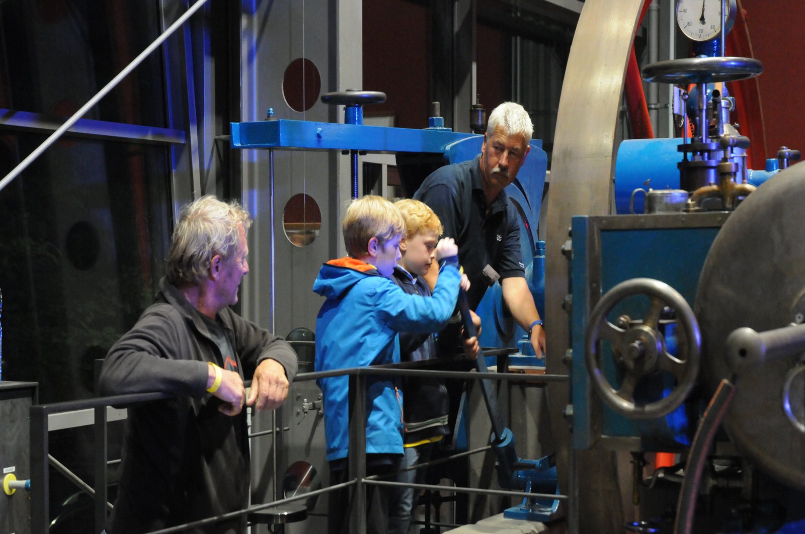 Zwei Jungen dürfen unter Aufsicht zwei erfahrener Männer einen Hebel an einer großen Maschine bedienen.