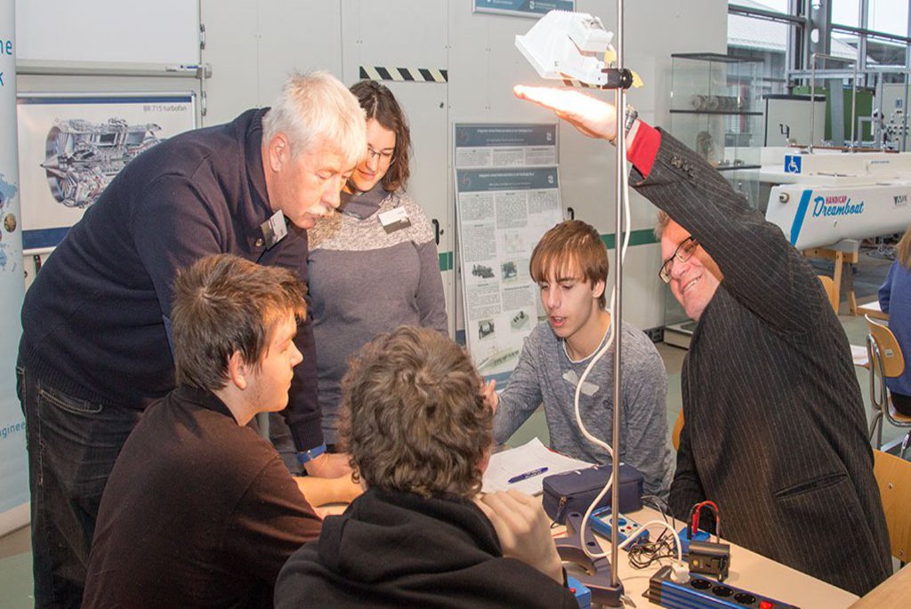 Studierende sitzen vor einem Versuchsaufbau und werden durch einen Professor unterstützt.