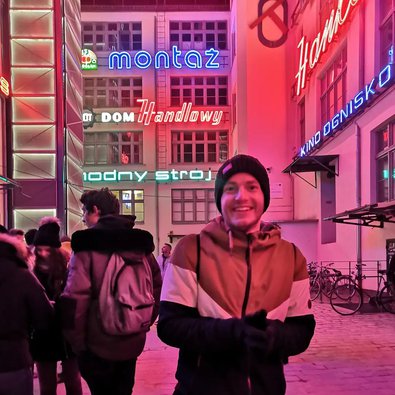Frederik grinst breit und steht auf dem Weihnachtsmarkt. Er ist dick eingepackt und hat die Hände in Handschuhen und eine warme Mütze auf