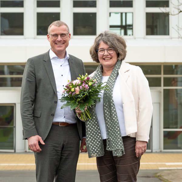 Ein Mann (Prof. Björn Christensen) und eine Frau (Prof. Ruth Boerckel) stehen nebeneinander vor einem Gebäude. Ruth Boerckel hält einen Blumenstrauß in der Hand.
