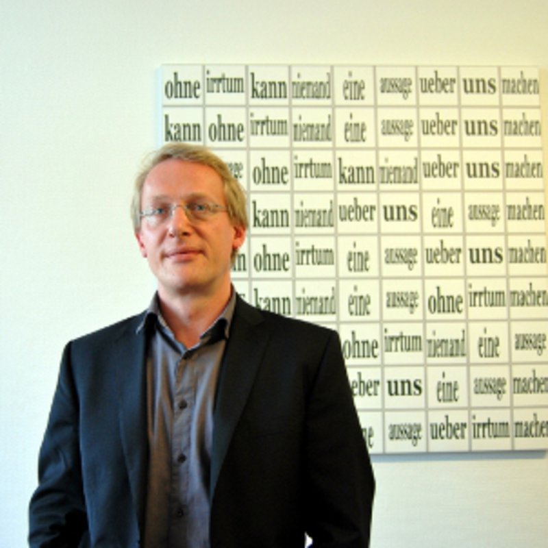 Ein Mann in schwarzem Jackett, steht vor einem Bild, auf dem ein Zitat gesschrieben steht.