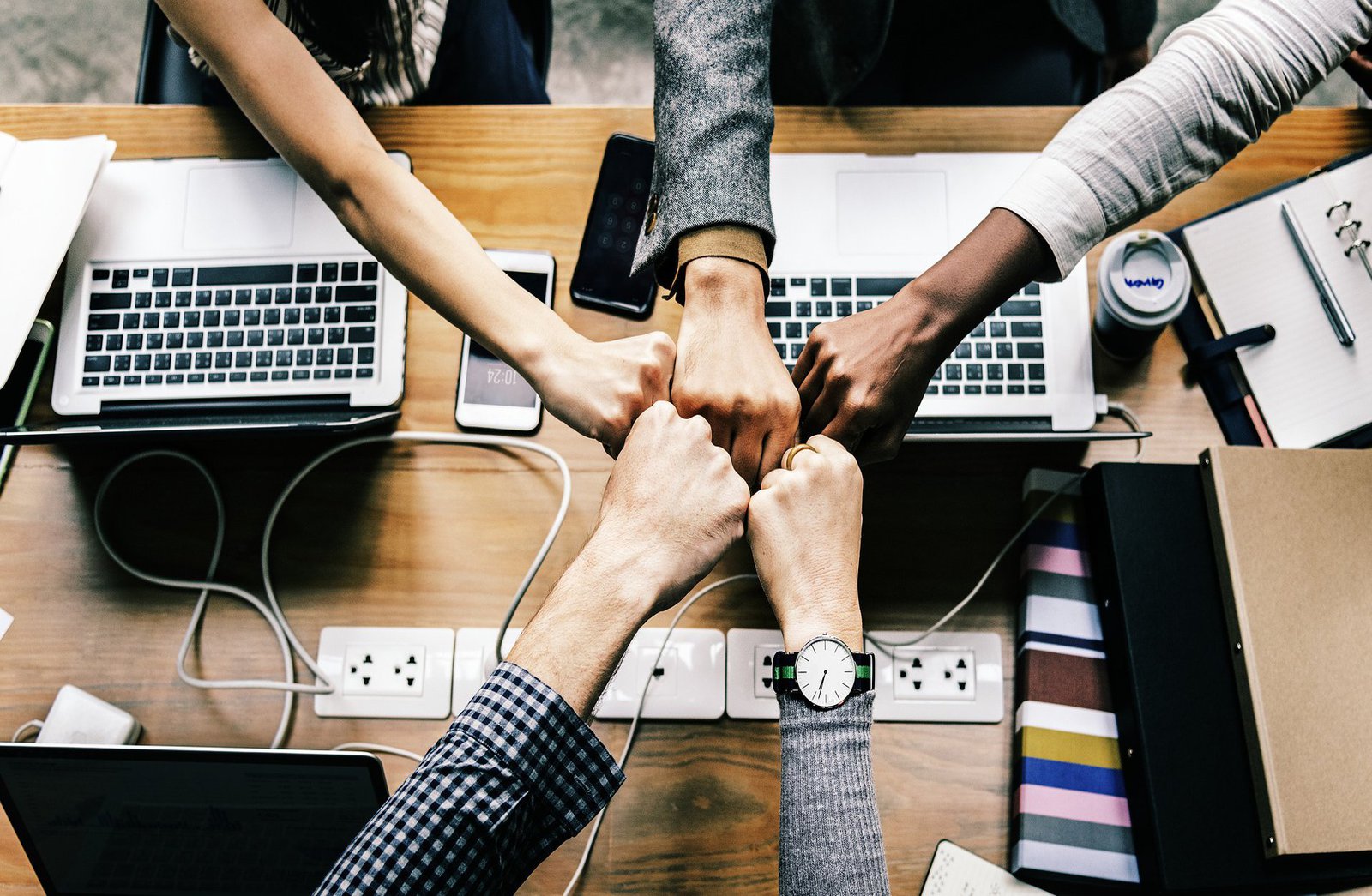 Fünf Fäuste treffen über einem mit Laptops und Handys beladenen Schreibtisch aufeinander.
