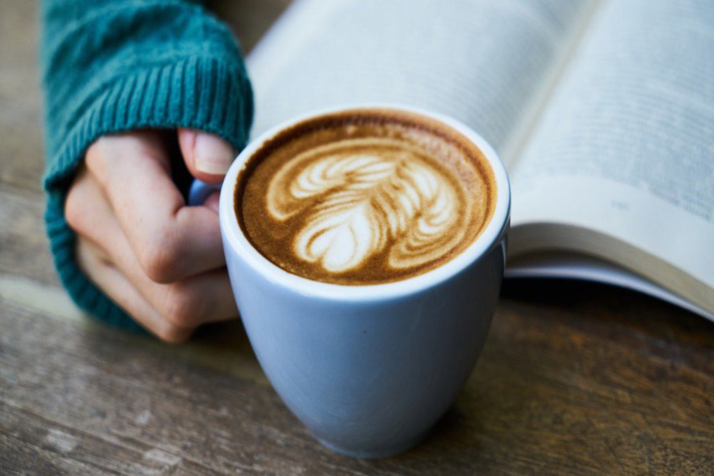 Eine Hand hält einen Cappucino, der neben einem aufgeschlagenen Buch steht.