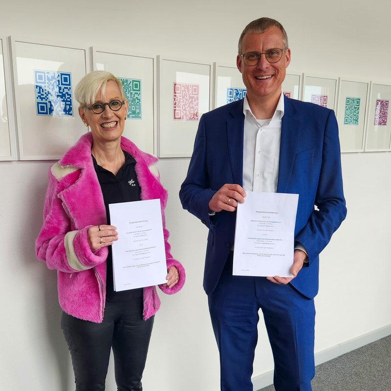 Eine Frau und ein Mann stehen nebeneinander, halten ein Stück Papier in die Kamera und lächeln.   