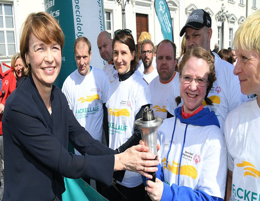 Eine Frau, in schwarzer Bluse und kariertem Rock, überreicht einem Laufteam eine Fackel.