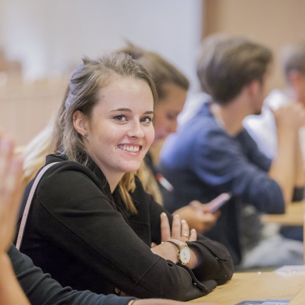 Seitlichere Blick in einen Stuhlreihe im Hörsaal, eine junge Frau lächelt in die Kamera
