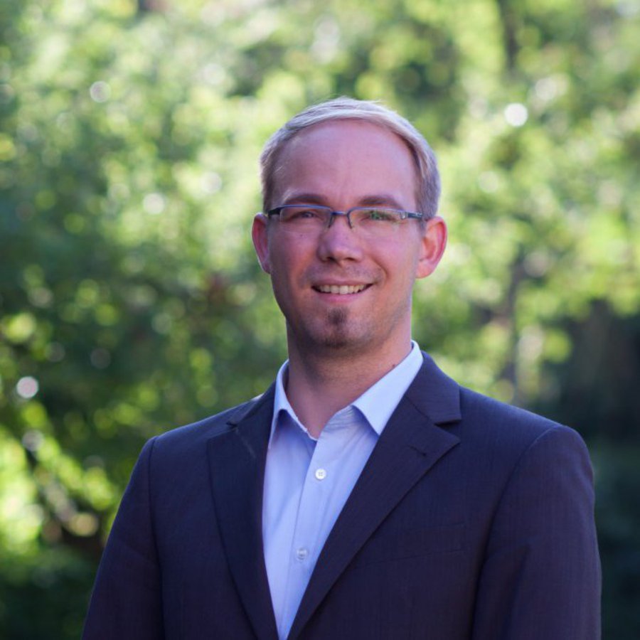 Ein Mann in helllblauem Hemd und dunkelblauem Sakko posiert im freien.