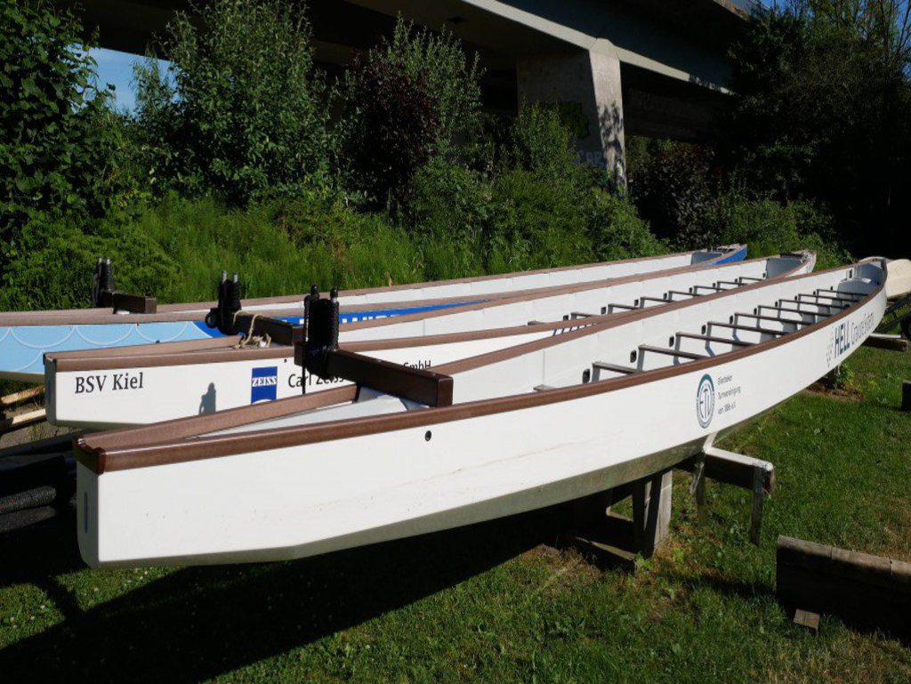 Drei Ruderboote stehen auf Böcken nebeneinander. Im Hintergrund ist eine Brücke zu sehen.