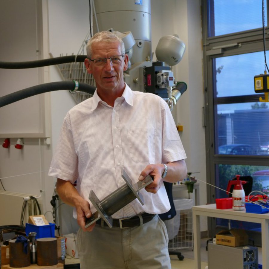 Ein Mann in weissem Hemd steht in einem Maschinenraum und hält ein Werkstück aus Metall in den Händen.