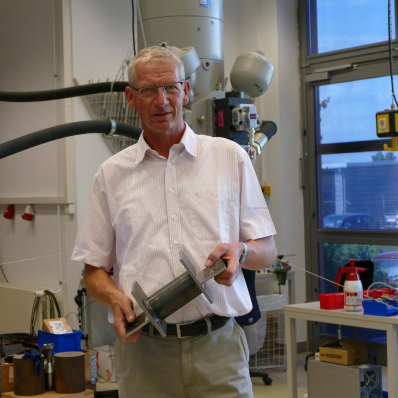 Ein Mann in weissem Hemd steht in einem Maschinenraum und hält ein Werkstück aus Metall in den Händen.