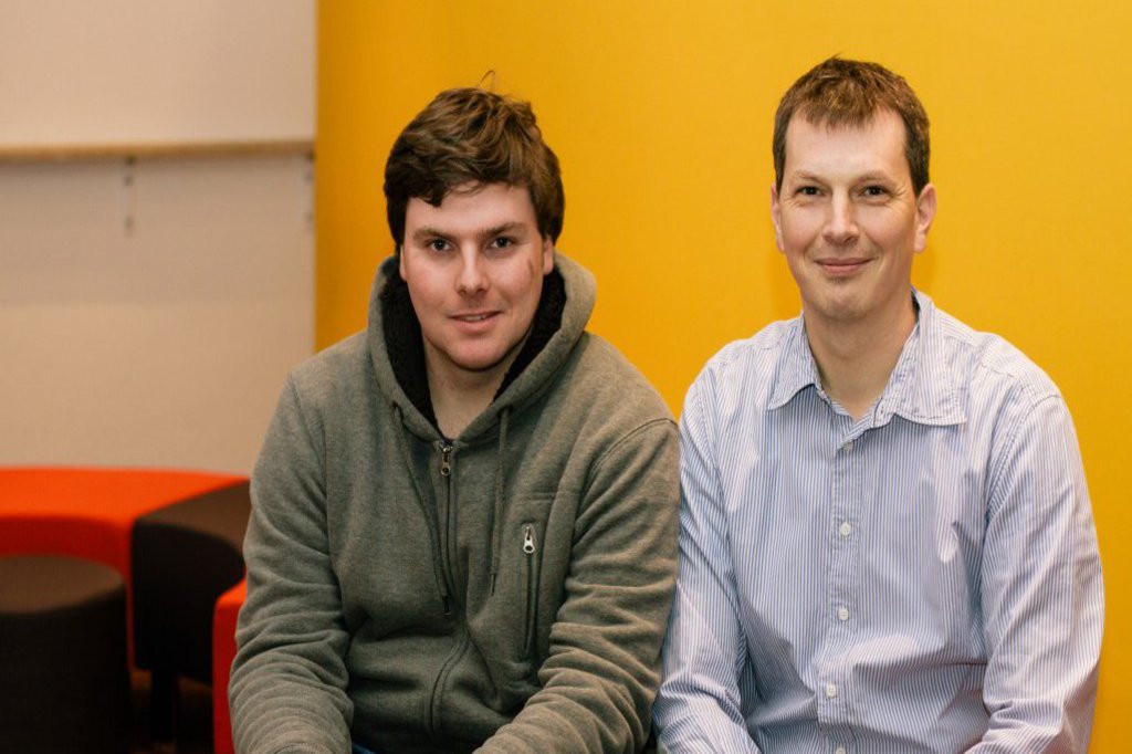 Zwei nebeneiander sitzende Männer, grinsen vor einer gelben Wand in die  Kamera.