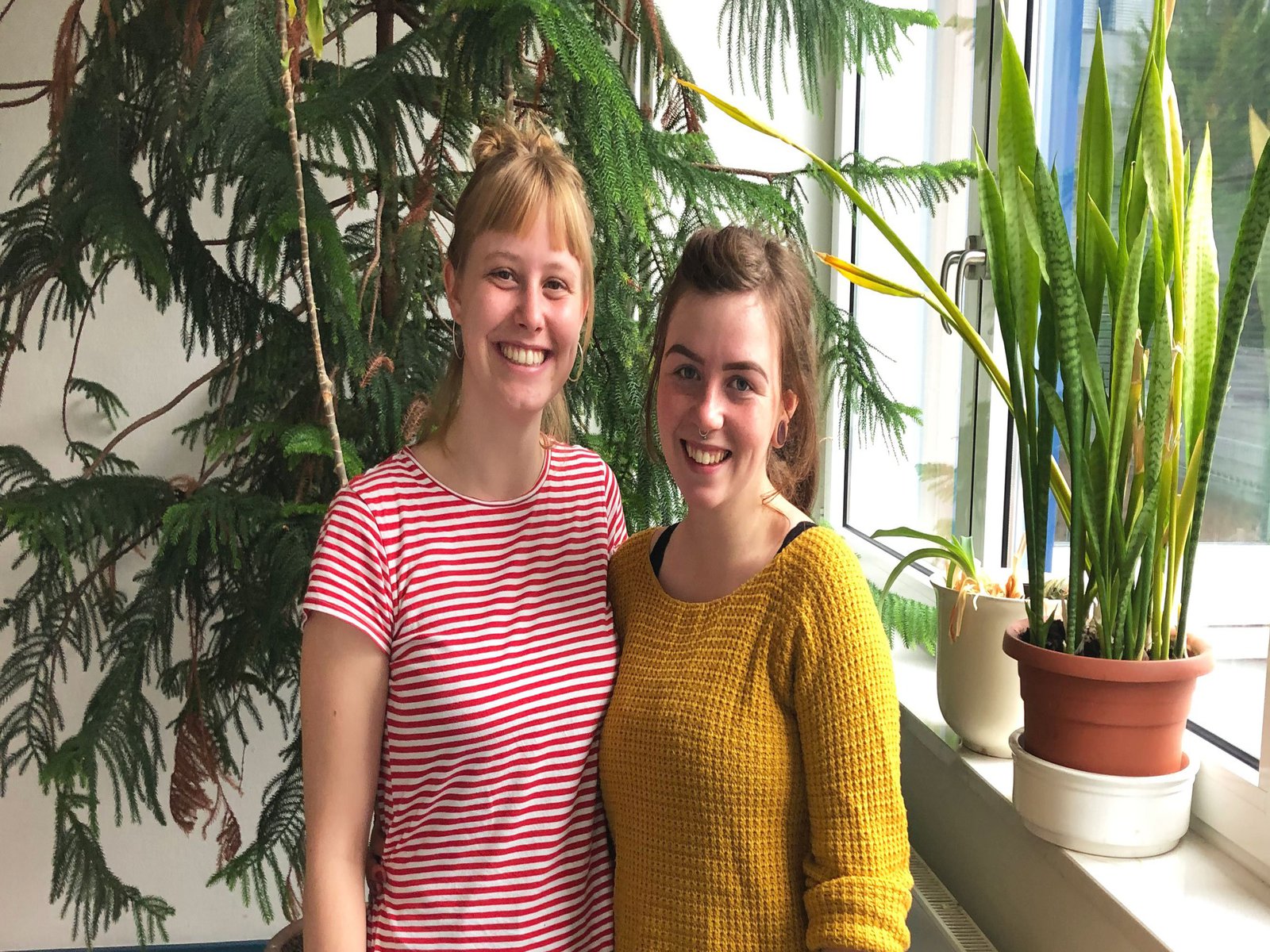 Zwei Frauen in bunten T-Shirts posieren vor einer Wand mit hohen grünen Zimmerpflanzen.