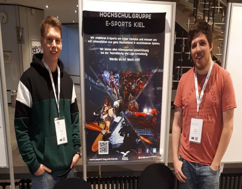 Zwei e-Sportler stehen vor dem Werbeplakat für die Hoschulgruppe e-Sports Kiel.