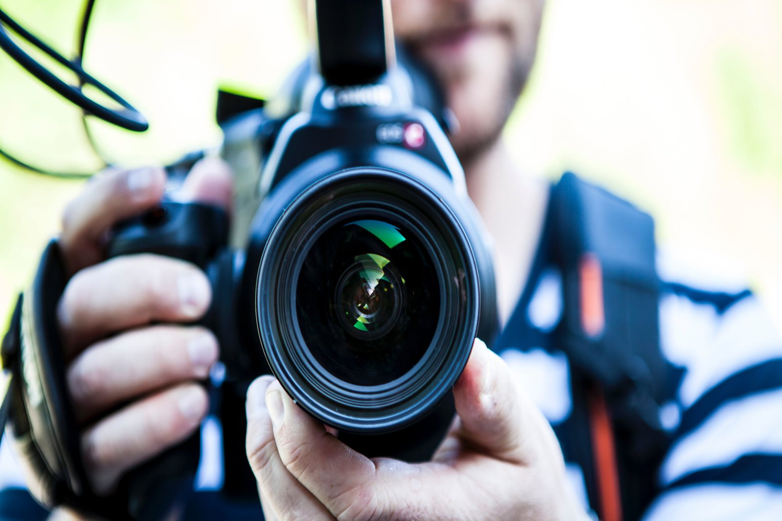 Der Fokus liegt auf der Linse einer Kamera. Im Hintergrund steht ein Mann, der diese hält.