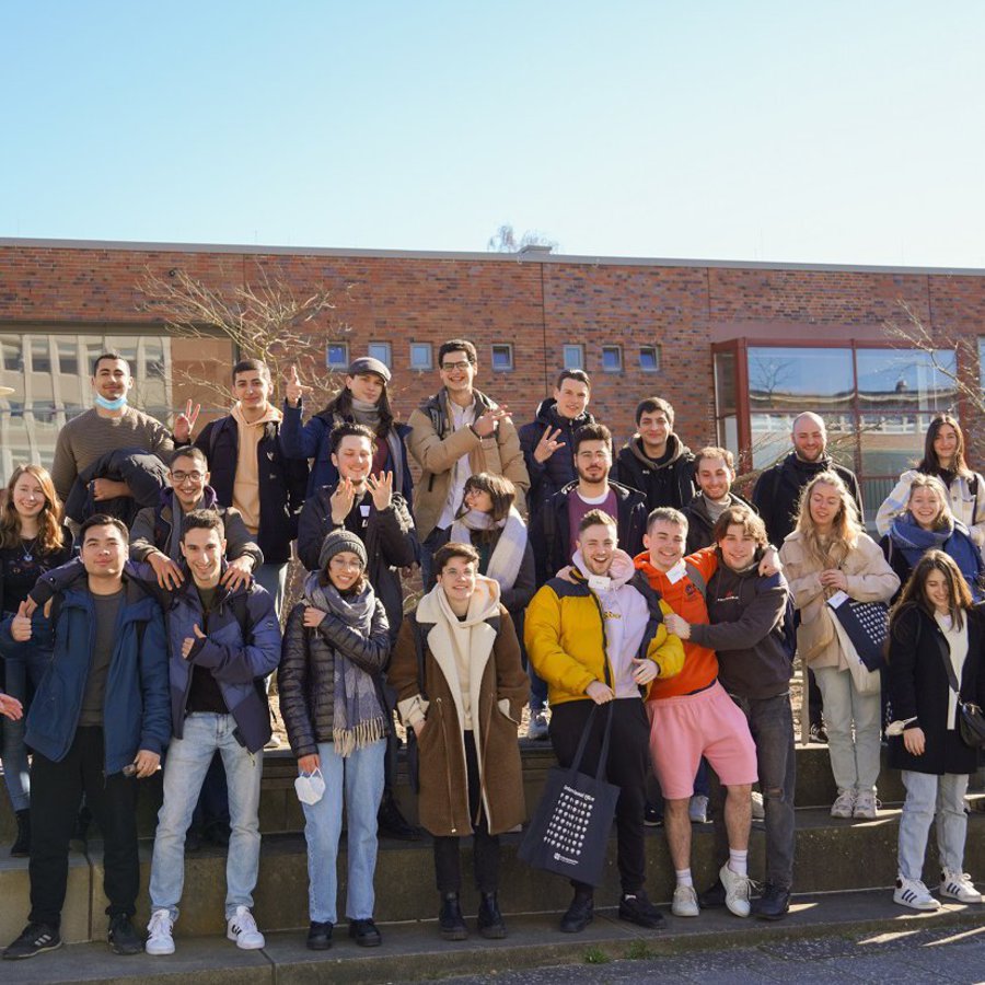 Eine große Gruppe junger Frauen und Männer steht aufgereiht auf drei Stufen und winkt und lacht.