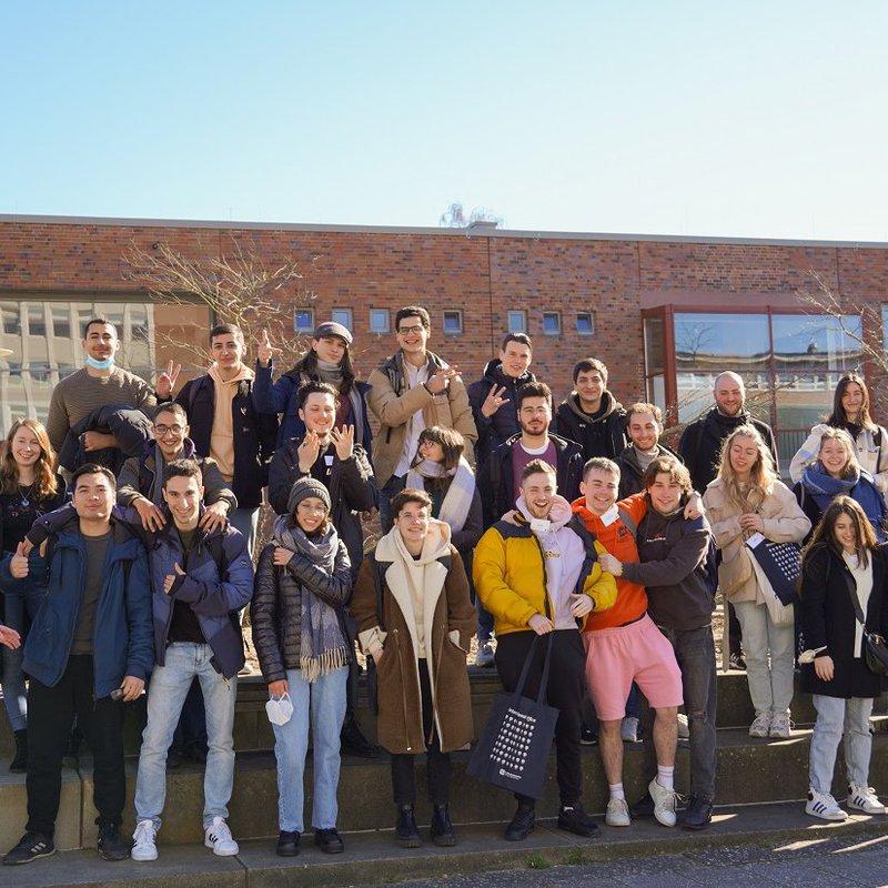 Eine große Gruppe junger Frauen und Männer steht aufgereiht auf drei Stufen und winkt und lacht.