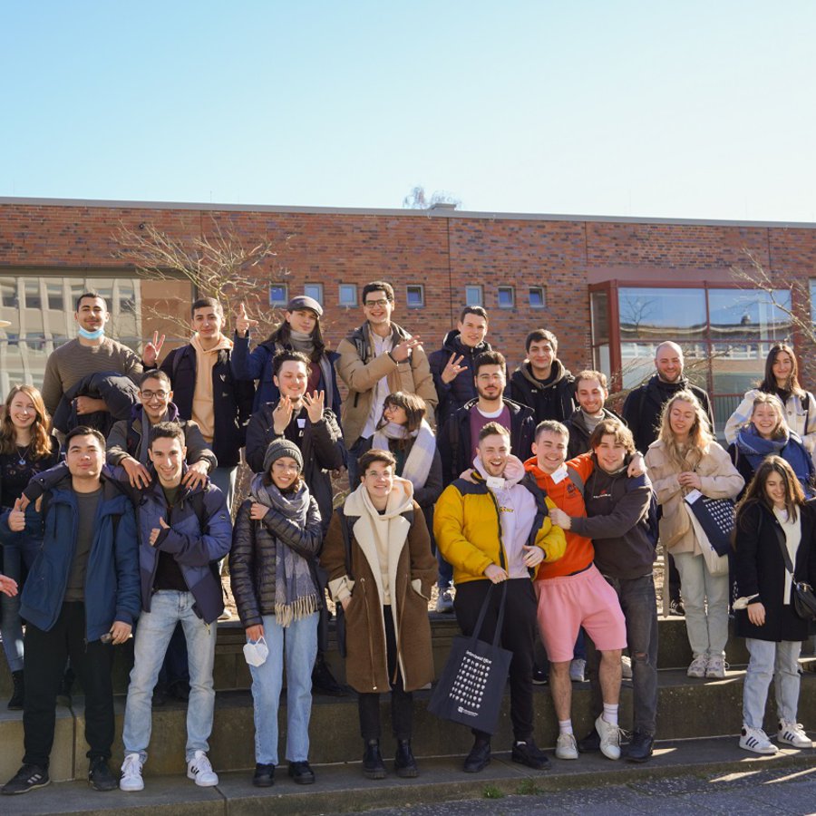 Eine große Gruppe junger Frauen und Männer steht aufgereiht auf drei Stufen und winkt und lacht.