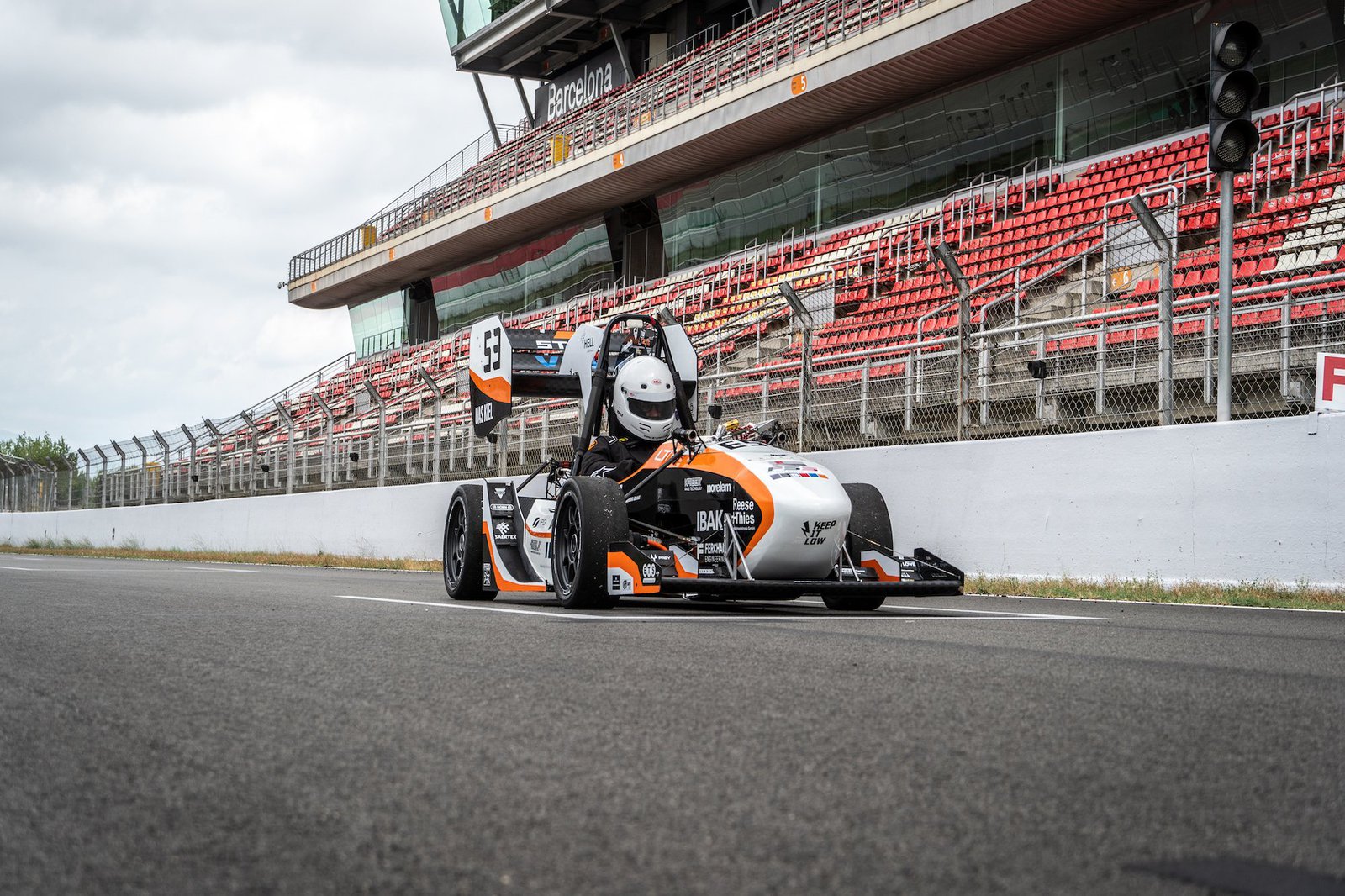 Ein bemannter weiss,schwarz, orangener Rennwagen steht alleine auf einer Rennstrecke. Die im Hintergrund zu sehende Tribüne ist menschenleer.
