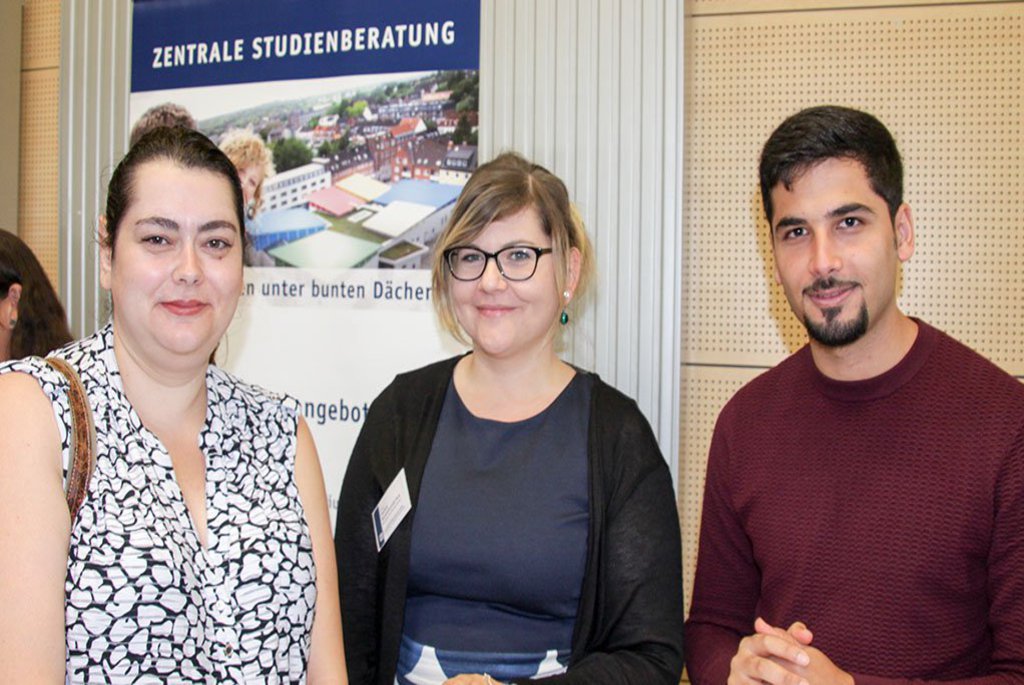Zwei Frauen und eine Mann grinsen an einem Infostand in die Kamera.