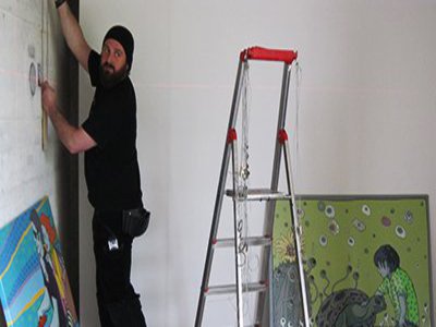 Ein Mann in schwarzer Kleidung ist mit einer Leiter ausgestattet und bringt Bilder an.