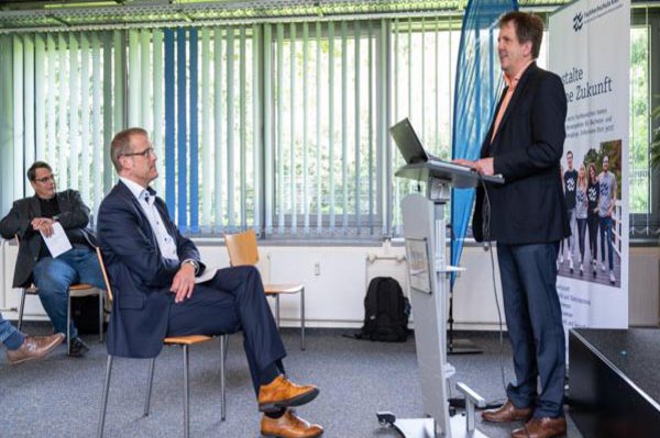 drei Männer, einer spricht vom Podium ins Auditorium