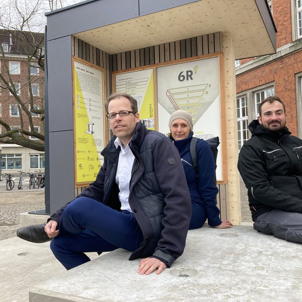 Zwei Männer und eine Frau sitzen auf einem Betonblock und schauen in die Kamera. Im Hintergrund sind Infotafeln zu sehen.