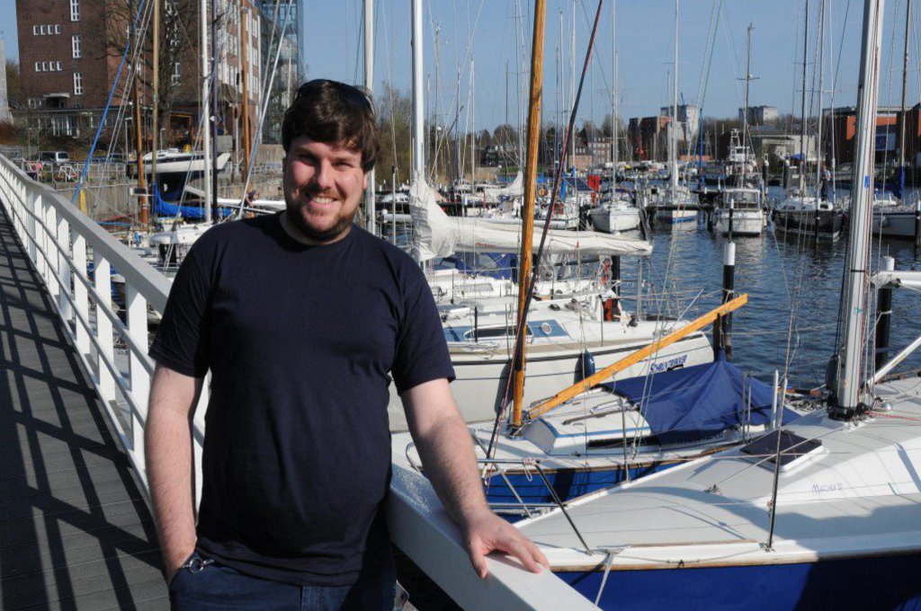 Ein Mann steht am Bootssteg, eines Yachthafens und lächelt in die Kamera.