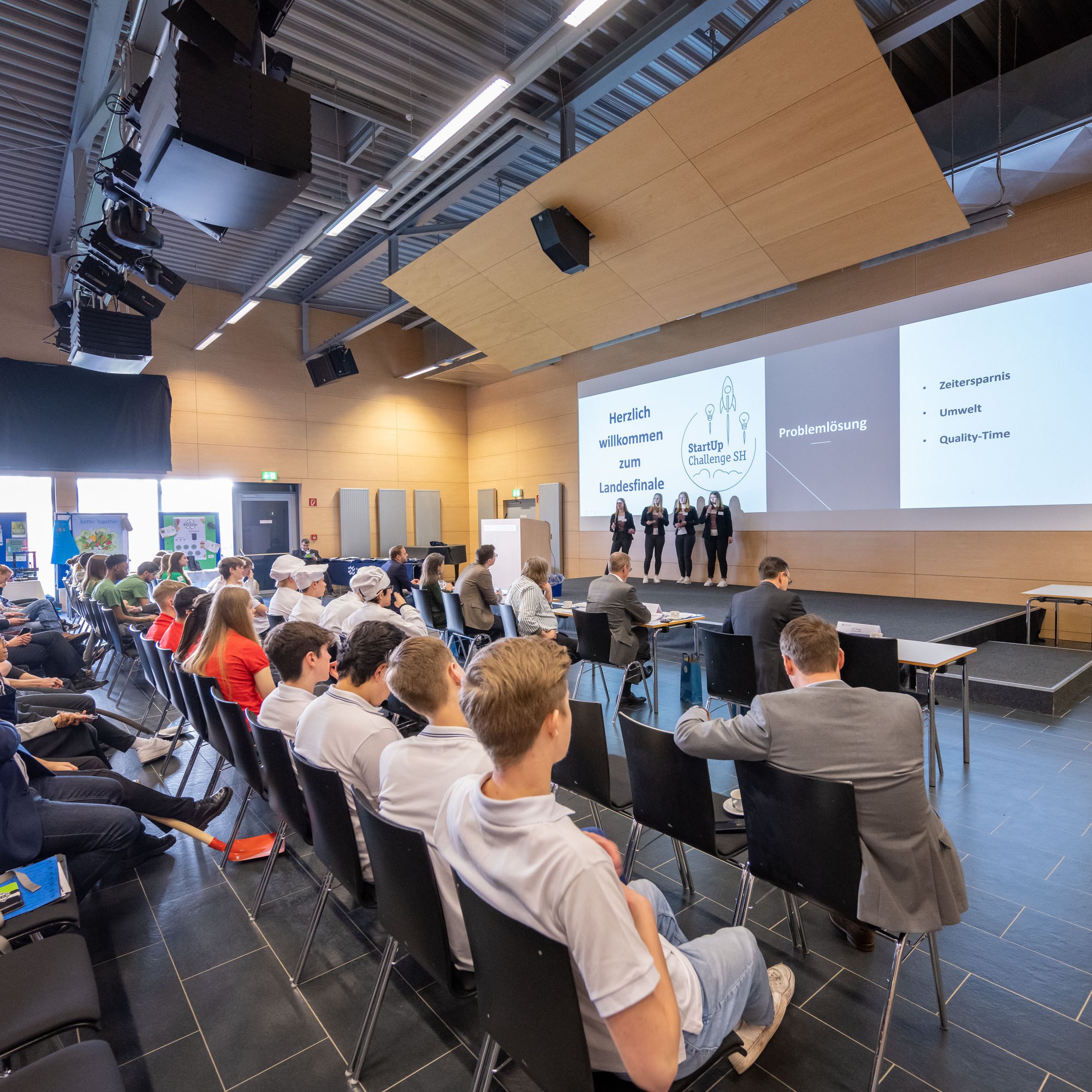 StartUp Challenge SH Landesfinale In einem großen Raum stehen vier junge Menschen auf der Bühne vor Publikum