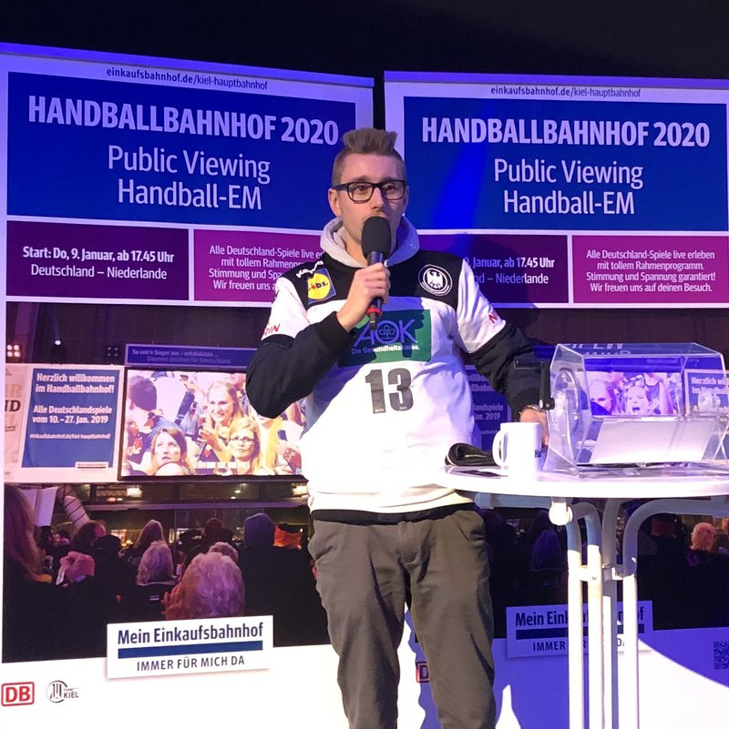 Mann mit Mikro vor Aufsteller mit Handballbahnhof-Schriftzug