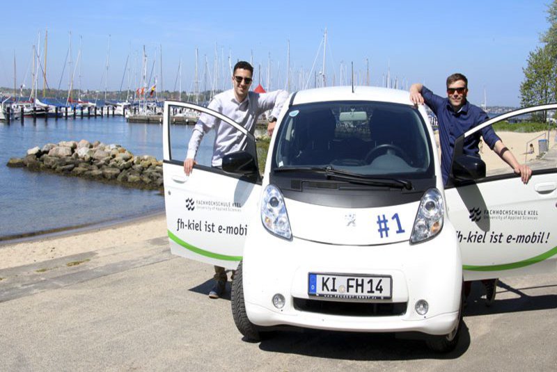 Zwei Männer stehen bei geöffneten Türen an einem E-Auto. Im Hintergrund ist ein Strandabschnitt zu sehen.