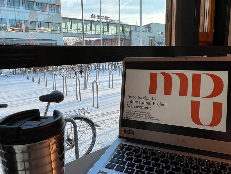 ein aufgeklappter Laptop mit Vorlesungsfolien, daneben ein Thermosbecher mit Heißgetränk. Durch große Glasfenster sieht man den Campus