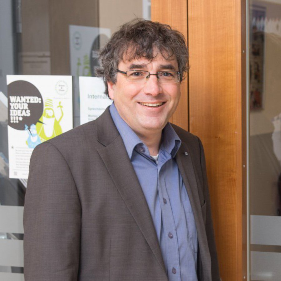 Ein Mann in grauem Sakko und blauem Hemd steht lächelnd an der eingangstür des International Office der FH Kiel und lächelt freundlich in die Kamera.