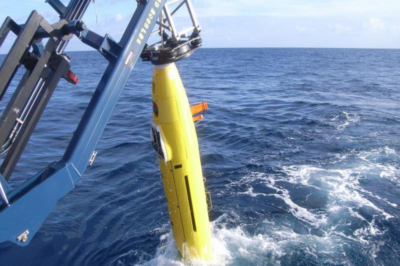 Mitten im Meer wird ein gelbes torpedoartiges Geraät, mit einem Kran, aus dem Wasser geholt.