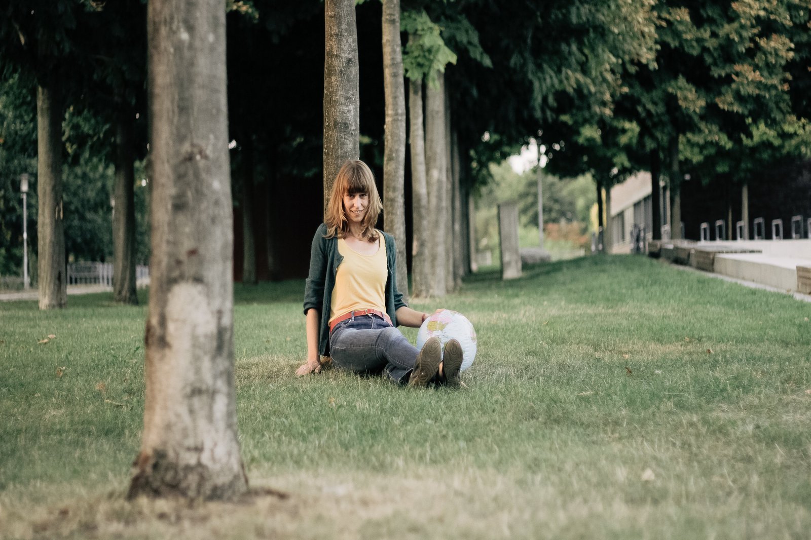 Eine Frau sitzt auf einem gepflegten Rasen und leht sich an einen dünnen Baum an. Neben ihr stützt ihre Hand auf einem aufblasbaren Globus.