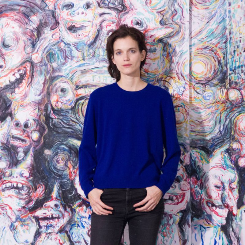 Eine Frau in blauem Pullover posiert vor einem bizarren Gemälde.
