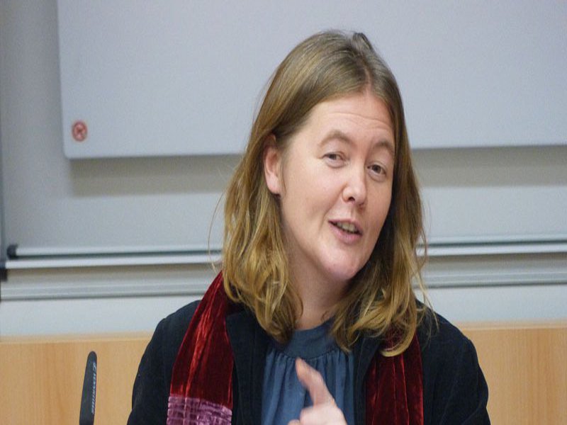 Eine Frau hat einen roten Schal um den Hals geworfen und spricht mit erhobenem Finger zu ihren Zuhörenden.