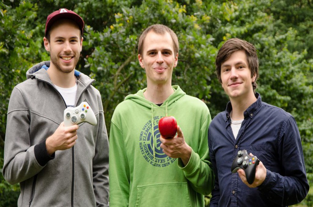 Drei Männer stehen nebeneinander im Freien. Zwei von ihnen halten Controller und einer einen roten Apfel in der Hand.