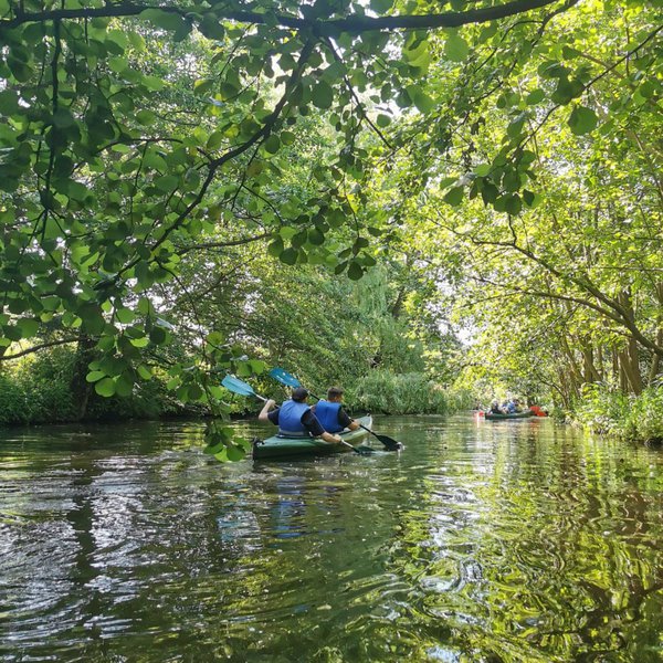 Zwei Personen in einem Kanu auf einem Fluss unter Bäumen.