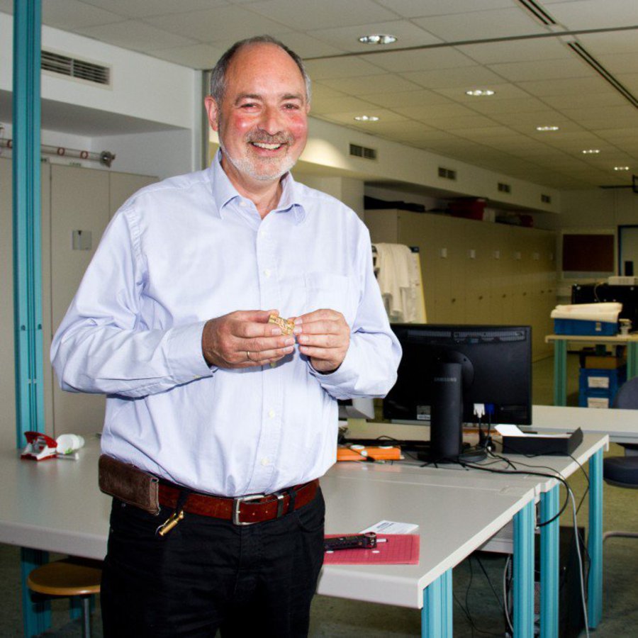 Ein gut gelaunter älterer Mann, mit Ledergürtel, steht in einem Labor.