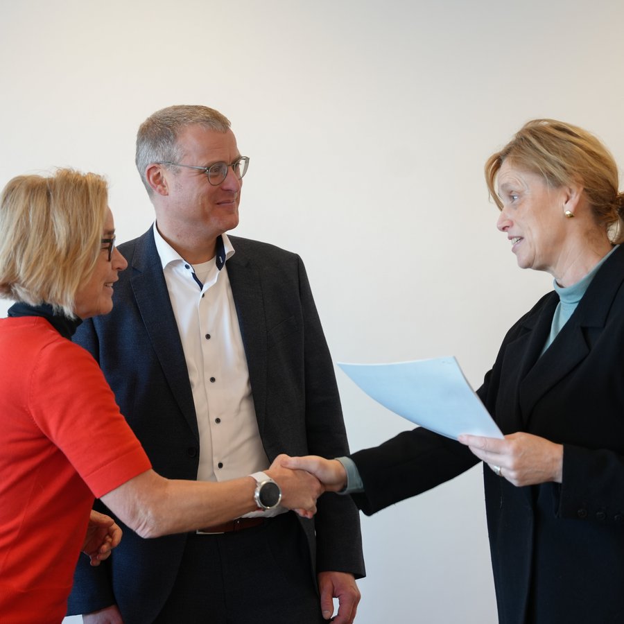 Auf dem Bild schüttelt Christiane Hipp (Präsidentin der EUF) Ministerin Karin Prien (MBWFK) die Hand. Björn Christensen steht dahinter. Karin Prien hält ein Papier in der Hand. 