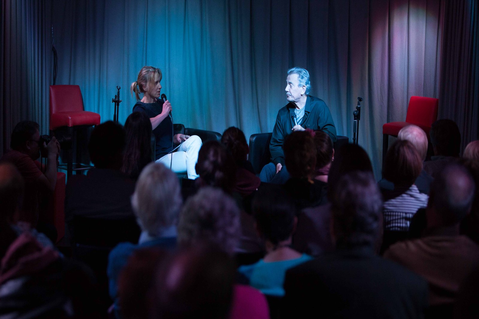 Eine dezent belichtete Bühne, auf der eine Frau und ein älterer Mann sitzen.