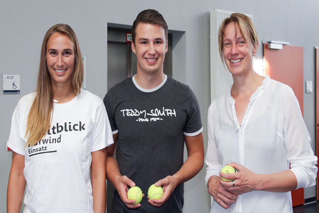 Zwei Frauen und eine Mann posieren mit Tennisbällen in den Händen auf einem Flur.