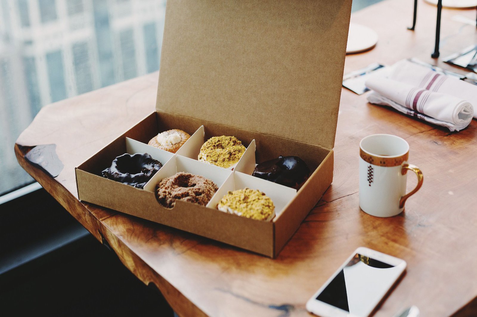 Eine Pappschachtel ist mit sechs Donuts befüllt und liegt auf einem Holztisch. Nebendran liegt ein Telefon und eine Kaffeetasse.