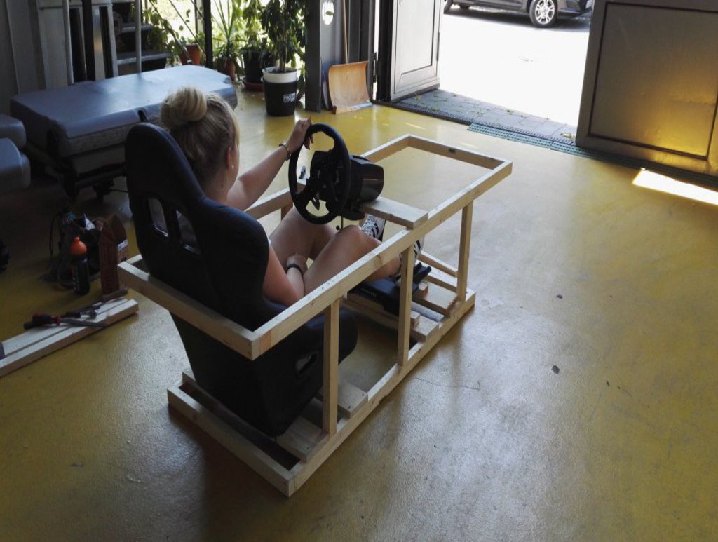 Eine Frau sitzt in einer Holzkonstruktion mit Schalensitz, Lenkrad und Pedalen.