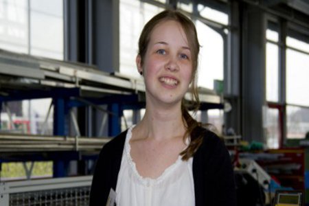 Eine Studentin steht an Metallregalen und schaut freundlich in die Kamera.