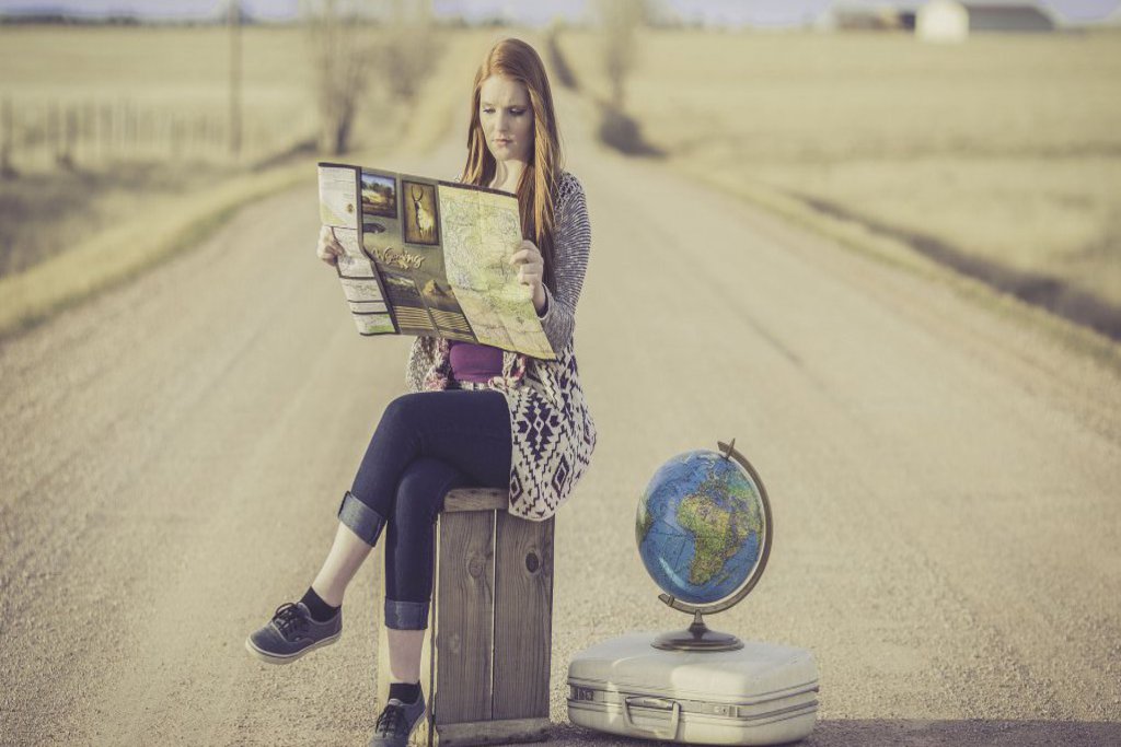 Eine Frau sitzt auf einer Kiste, in der Mitte einer Landstraße und liest eine Karte.