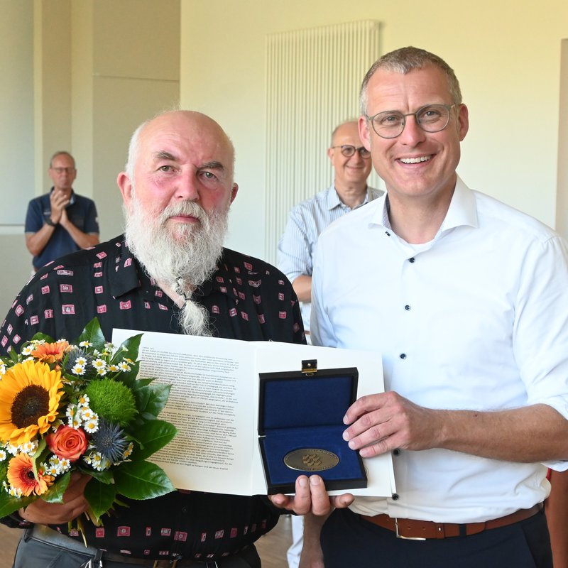 Ein Mann mit Blumenstrauß und Urkunde, die er von einem anderen Mann überreicht bekommt. Im Hintergrund applaudieren stehende Personen.