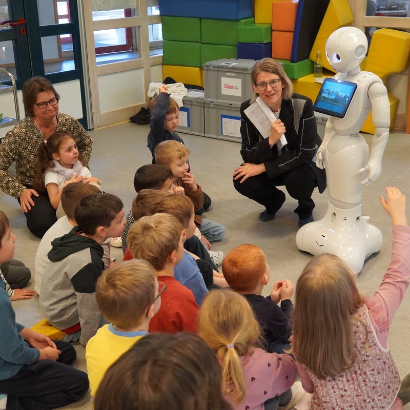 Frau kniet neben Roboter vor Kindern