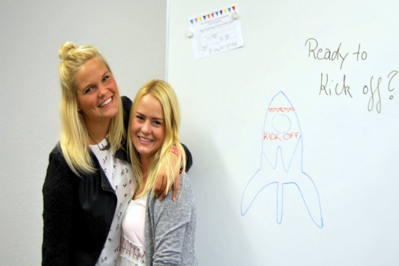 Zwei Frauen stehen Schulter an Schulter neben einem Whiteboard, auf dem eine Rakete zu sehen ist.